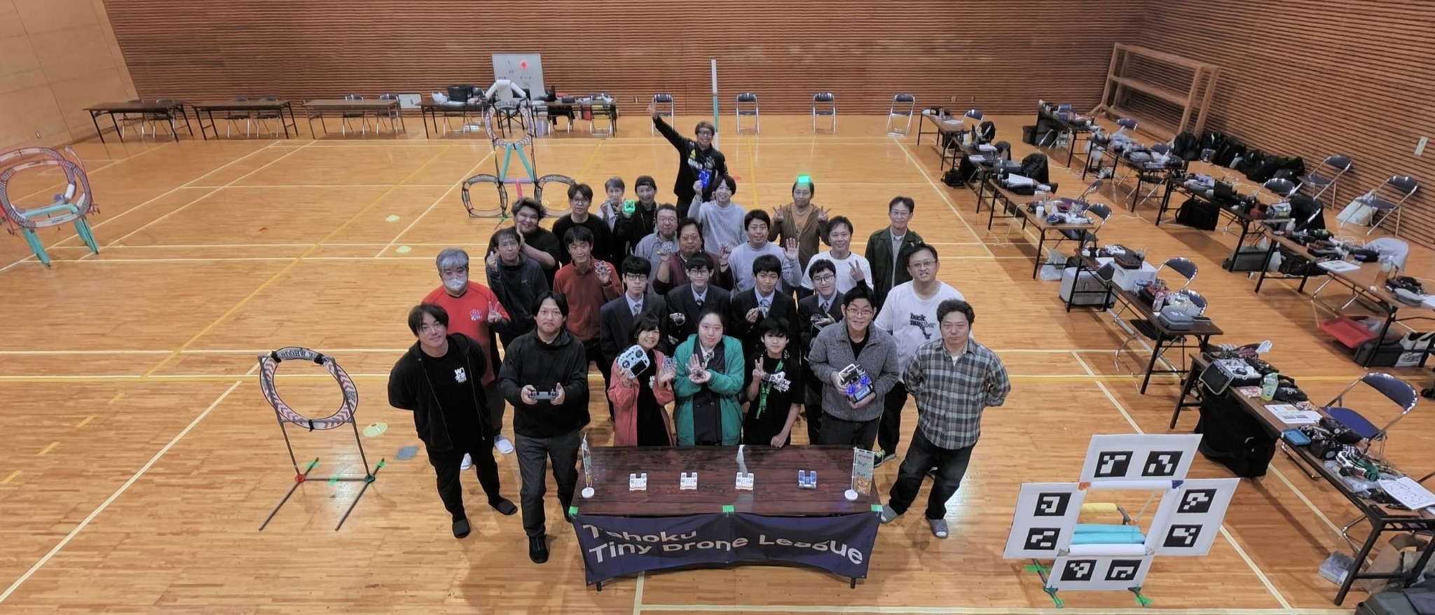 WTW秋田予選の集合写真。参加者の周囲にTinyWhoopのコースや機材が並ぶ会場